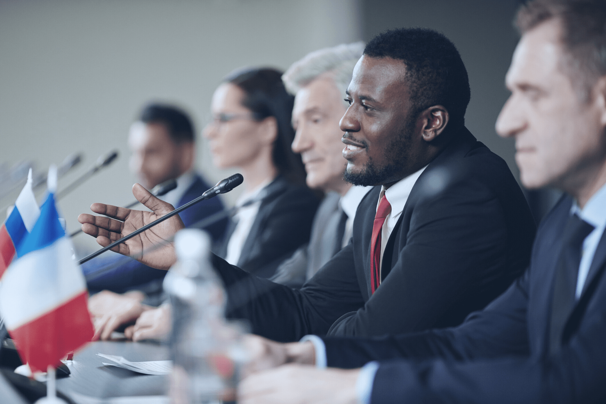 Various people in suits sit at a table in the style of a panel discussion. There are microphones and various country flags before them.
