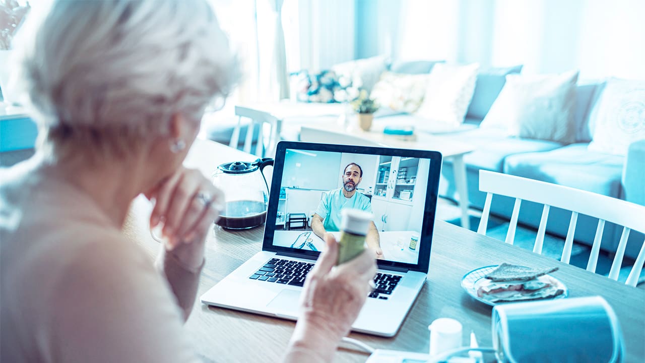 An elderly person holding a prescription bottle while in teleconference with a doctor.